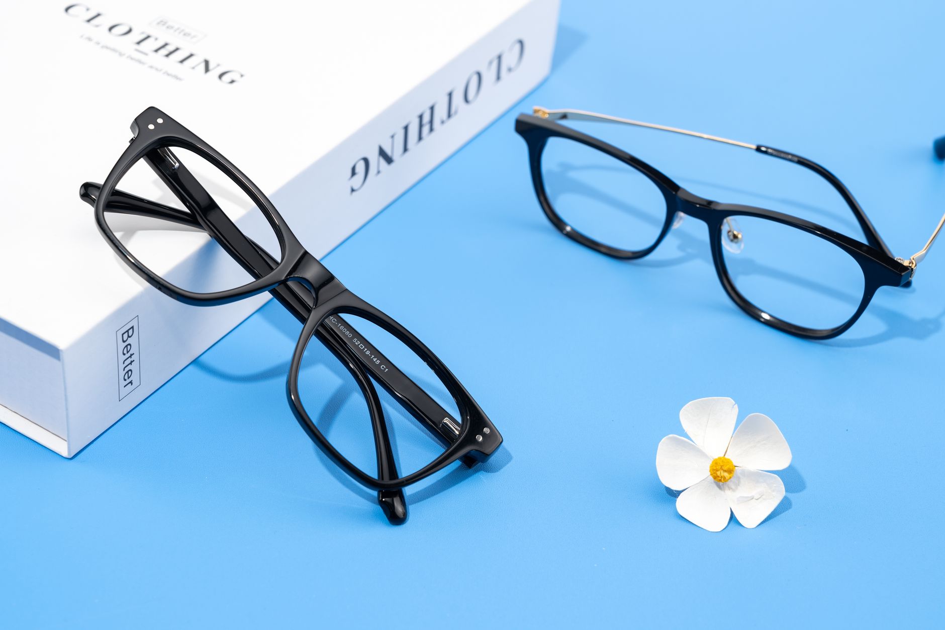Minimalist display of black eyeglasses on blue background with a white flower and cloth box.